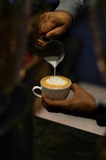 A person is carefully pouring milk into a cup of coffee to create latte art. The focus is on the intricate design forming on the surface of the coffee, suggesting a skilled barista at work.