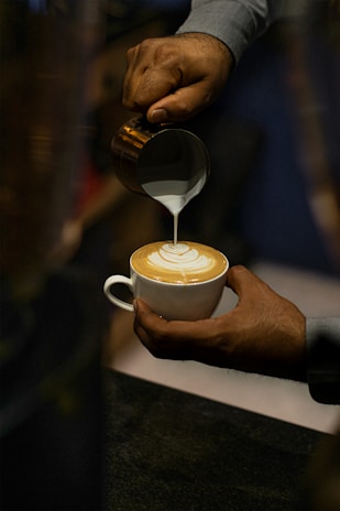 A person is carefully pouring milk into a cup of coffee to create latte art. The focus is on the intricate design forming on the surface of the coffee, suggesting a skilled barista at work.