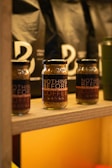 Several jars labeled 'Nothing Before Hazelnut Instant Coffee' are arranged on a wooden shelf. Behind them, there are black bags with minimalistic white designs. The setting appears to be a rustic or artisanal store or cafe. The lighting is warm, enhancing the cozy and inviting atmosphere.