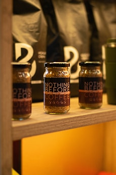 Several jars labeled 'Nothing Before Hazelnut Instant Coffee' are arranged on a wooden shelf. Behind them, there are black bags with minimalistic white designs. The setting appears to be a rustic or artisanal store or cafe. The lighting is warm, enhancing the cozy and inviting atmosphere.
