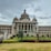 A grand, ornate building with a central dome and two side domes, featuring classical architectural elements like columns and intricate carvings. An Indian flag flies atop the central dome, and the facade bears the inscription 'Government Work is God's Work'. Lush greenery surrounds the structure, with hedges and manicured lawns in the foreground. The sky appears overcast, suggesting an impending storm or recent rain.