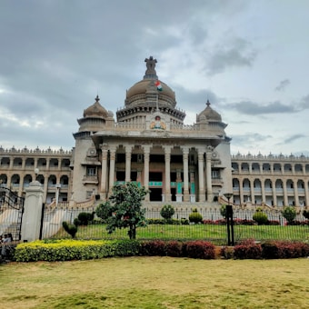 A grand, ornate building with a central dome and two side domes, featuring classical architectural elements like columns and intricate carvings. An Indian flag flies atop the central dome, and the facade bears the inscription 'Government Work is God's Work'. Lush greenery surrounds the structure, with hedges and manicured lawns in the foreground. The sky appears overcast, suggesting an impending storm or recent rain.