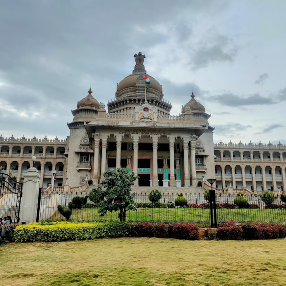 A grand, ornate building with a central dome and two side domes, featuring classical architectural elements like columns and intricate carvings. An Indian flag flies atop the central dome, and the facade bears the inscription 'Government Work is God's Work'. Lush greenery surrounds the structure, with hedges and manicured lawns in the foreground. The sky appears overcast, suggesting an impending storm or recent rain.