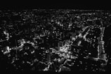 A cityscape at night with streets and buildings illuminated by artificial lights. The image presents a vast network of roads and high-rise structures, with a shimmering pattern of lights creating a vibrant urban scene.