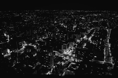 A cityscape at night with streets and buildings illuminated by artificial lights. The image presents a vast network of roads and high-rise structures, with a shimmering pattern of lights creating a vibrant urban scene.