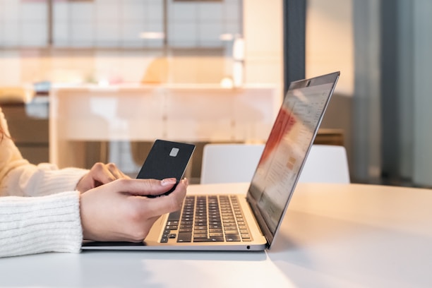 A person wearing a white sweater is sitting at a table and holding a credit card while using a laptop. The setting appears to be indoors with a modern and minimalistic design, featuring a bright and slightly blurred background.