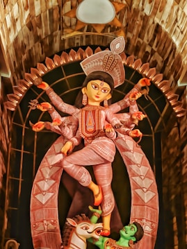 A statue of a multi-armed goddess adorned with intricate patterns and vibrant colors. The figure is posed gracefully, with one leg raised and appears to be standing atop a defeated figure. The setting includes a decorative arch and is richly detailed.