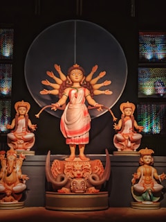 A depiction of a divine figure with multiple arms, wearing a traditional saree, standing on a detailed pedestal with intricate designs. Smaller deities accompany the central figure, each seated in a meditative pose. The setting includes ornate and colorful decorations in the background.