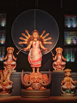 A depiction of a divine figure with multiple arms, wearing a traditional saree, standing on a detailed pedestal with intricate designs. Smaller deities accompany the central figure, each seated in a meditative pose. The setting includes ornate and colorful decorations in the background.