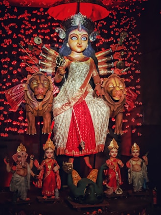 A vibrant depiction of a multi-armed goddess adorned in a red and white sari, sitting atop two large lions. The figure is surrounded by smaller statues, possibly other deities, each intricately dressed and positioned. The background is filled with glowing red lights, and the scene is set under a red canopy, emphasizing a festive or ritualistic atmosphere.