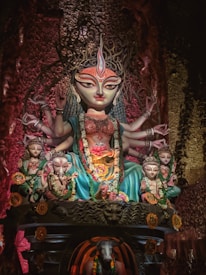 A colorful and intricate statue of a multi-armed goddess, adorned with a crown and surrounded by smaller figures. The goddess is wearing ornate jewelry and flowing garments. Festive garlands decorate the scene.