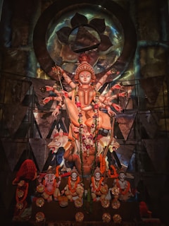 A richly decorated statue of a multi-armed deity, adorned with intricate garlands and ornaments. The statue is set against an ornate backdrop with geometric patterns and metallic accents, symbolizing spiritual and cultural significance.