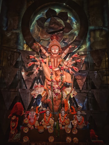 A richly decorated statue of a multi-armed deity, adorned with intricate garlands and ornaments. The statue is set against an ornate backdrop with geometric patterns and metallic accents, symbolizing spiritual and cultural significance.