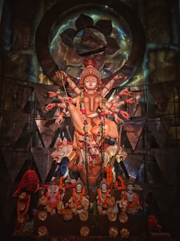 A richly decorated statue of a multi-armed deity, adorned with intricate garlands and ornaments. The statue is set against an ornate backdrop with geometric patterns and metallic accents, symbolizing spiritual and cultural significance.