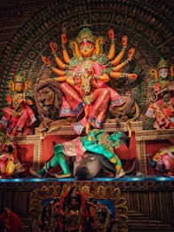 An elaborate and colorful depiction of a Hindu goddess, adorned with multiple arms and intricate jewelry, mounted on a lion and defeating a demon underfoot. The background is richly decorated with patterns and other figures also showcased in vibrant attire and accessories.