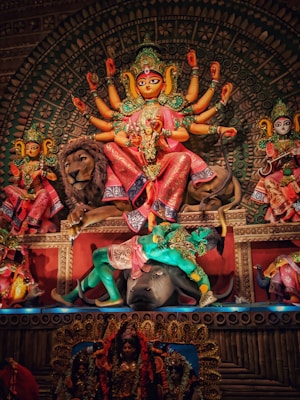 An elaborate and colorful depiction of a Hindu goddess, adorned with multiple arms and intricate jewelry, mounted on a lion and defeating a demon underfoot. The background is richly decorated with patterns and other figures also showcased in vibrant attire and accessories.
