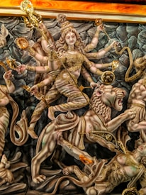 A highly detailed and ornate depiction of a multi-limbed goddess figure adorned in golden attire and accessories, shown riding a lion. The goddess holds various symbolic objects in her multiple arms, including a trident and a discus. The background features grayish, rock-like formations.