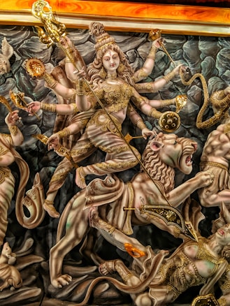 A highly detailed and ornate depiction of a multi-limbed goddess figure adorned in golden attire and accessories, shown riding a lion. The goddess holds various symbolic objects in her multiple arms, including a trident and a discus. The background features grayish, rock-like formations.