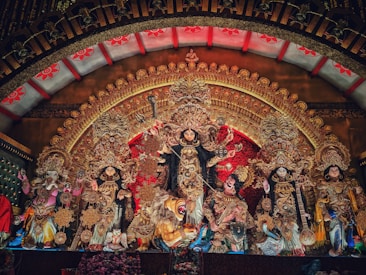 A vibrant and intricately detailed display of deities, featuring a central figure with multiple arms holding different objects, surrounded by other finely adorned figures. The backdrop is richly decorated with golden and red patterns, and there is a prominent lion figure at the bottom.