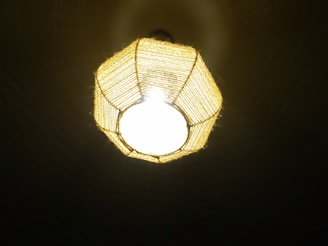 A ceiling light fixture with a woven, geometric-shaped lampshade. The lampshade is made of a natural material that allows bright light to emit through, casting subtle patterns on the surrounding surfaces.