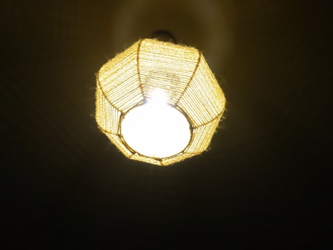 A ceiling light fixture with a woven, geometric-shaped lampshade. The lampshade is made of a natural material that allows bright light to emit through, casting subtle patterns on the surrounding surfaces.