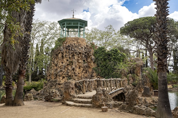 A natural landscape featuring a rocky structure with a wooden bridge leading up to it. A gazebo-like structure with a decorative top sits atop the rocky outcrop. The area is surrounded by lush greenery, including tall trees and palm trees, under a partly cloudy sky.