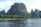 A scenic view of towering karst mountains covered in lush green vegetation, with a river flowing in the foreground. Small boats with people can be seen on the river, navigating the calm waters under a clear blue sky.