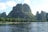 A scenic view of towering karst mountains covered in lush green vegetation, with a river flowing in the foreground. Small boats with people can be seen on the river, navigating the calm waters under a clear blue sky.