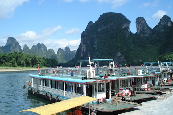 Several riverboats are docked along a calm river with large, green karst mountains in the background under a blue sky with scattered clouds. The boats have flat decks, lifebuoys, and appear to be set up for tourism or transport.