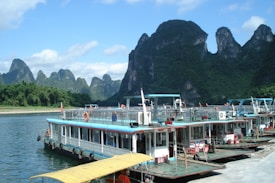 Several riverboats are docked along a calm river with large, green karst mountains in the background under a blue sky with scattered clouds. The boats have flat decks, lifebuoys, and appear to be set up for tourism or transport.