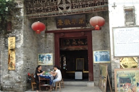 An ancient temple entrance with traditional Chinese architecture. Two large red lanterns hang above the doorway, flanked by historical and informational plaques. Two women are seated at a table outside, engaged in conversation.