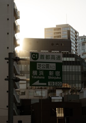 A cityscape with tall buildings, a prominent green highway sign featuring directions to Yokohama and Shinjuku, and the sun setting in the background casting a warm glow.