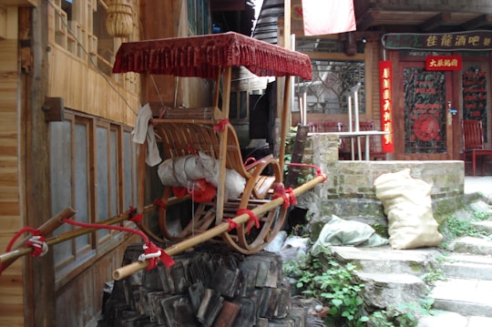 A wooden rickshaw with a red canopy is parked outside a traditional building. The scene includes a stack of roof tiles and bags by the stone steps. Chinese signage is visible on the building's facade, adding cultural elements to the setting.