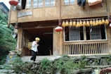 A traditional wooden house with a veranda featuring red lanterns and a string of dried corn hanging above. In front of the house, an elderly person wearing a yellow headscarf and carrying a basket is walking down stone steps. Lush greenery surrounds the building.