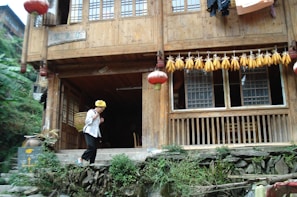 A traditional wooden house with a veranda featuring red lanterns and a string of dried corn hanging above. In front of the house, an elderly person wearing a yellow headscarf and carrying a basket is walking down stone steps. Lush greenery surrounds the building.