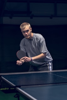 A person stands poised to hit a table tennis ball, holding a paddle in the ready position. The setting appears to be an indoor table tennis area with a dark background. The focus is on the player's intent concentration.