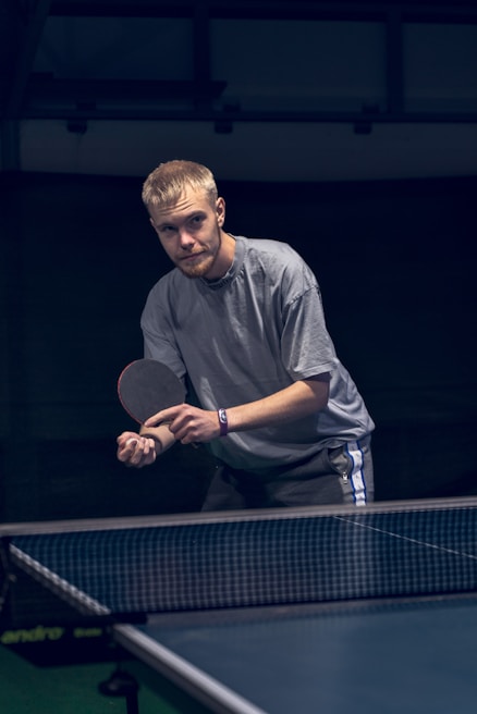 A person stands poised to hit a table tennis ball, holding a paddle in the ready position. The setting appears to be an indoor table tennis area with a dark background. The focus is on the player's intent concentration.