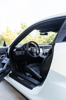 The interior view of a luxurious car featuring a sleek dashboard with a prominent steering wheel displaying a high-end logo. The seat is upholstered with premium materials, and the door is open, revealing intricate detailing and controls on the door panel. The car appears to be parked outdoors, with greenery visible in the background.