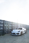 A sleek white sports car is parked on a gravel surface next to a row of gray shipping containers. The sunlight creates a bright, high-contrast setting, with the car's polished surface reflecting the light.