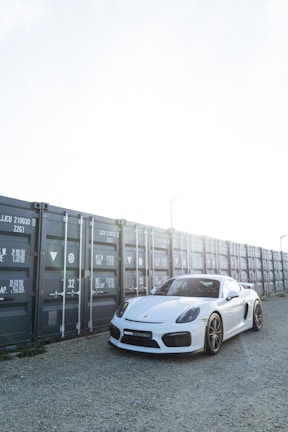 A sleek white sports car is parked on a gravel surface next to a row of gray shipping containers. The sunlight creates a bright, high-contrast setting, with the car's polished surface reflecting the light.