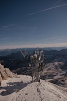 A telecommunications tower stands on a snow-covered mountain peak with rugged mountains and a clear blue sky in the background, creating a contrast between technology and nature.