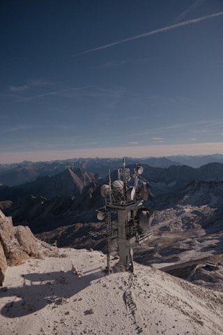 A telecommunications tower stands on a snow-covered mountain peak with rugged mountains and a clear blue sky in the background, creating a contrast between technology and nature.