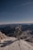 A telecommunications tower stands on a snow-covered mountain peak with rugged mountains and a clear blue sky in the background, creating a contrast between technology and nature.