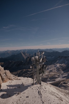 A telecommunications tower stands on a snow-covered mountain peak with rugged mountains and a clear blue sky in the background, creating a contrast between technology and nature.