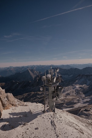A telecommunications tower stands on a snow-covered mountain peak with rugged mountains and a clear blue sky in the background, creating a contrast between technology and nature.