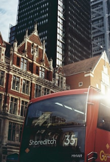 A red double-decker bus marked with the destination 'Shoreditch 35' is captured against the backdrop of modern skyscrapers and historic brick buildings. The bus is positioned in the lower right corner, while the buildings rise into the sky, merging old architectural styles with contemporary designs.
