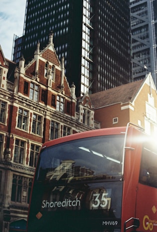 A red double-decker bus marked with the destination 'Shoreditch 35' is captured against the backdrop of modern skyscrapers and historic brick buildings. The bus is positioned in the lower right corner, while the buildings rise into the sky, merging old architectural styles with contemporary designs.