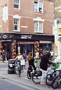 A bustling city street scene featuring several people interacting with rental bicycles in front of a brick building with a shop named 'Ensō Lounge'. The area is lively, with pedestrians walking nearby and the vibrant tiled facade of the shop adding color.
