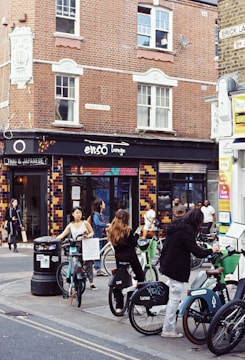 A bustling city street scene featuring several people interacting with rental bicycles in front of a brick building with a shop named 'Ensō Lounge'. The area is lively, with pedestrians walking nearby and the vibrant tiled facade of the shop adding color.