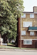 A residential scene with a brick apartment building featuring several windows, a green balcony, and a road sign that reads 'Dacre Park'. A tree with lush green foliage partially obscures the building. A street runs along the foreground, and a car is visible parked in the background. The atmosphere is quiet and suburban.